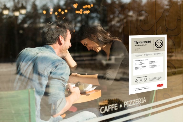 To gjester på et serveringssted med smilefjesresultat fra Mattilsynet på vinduet.